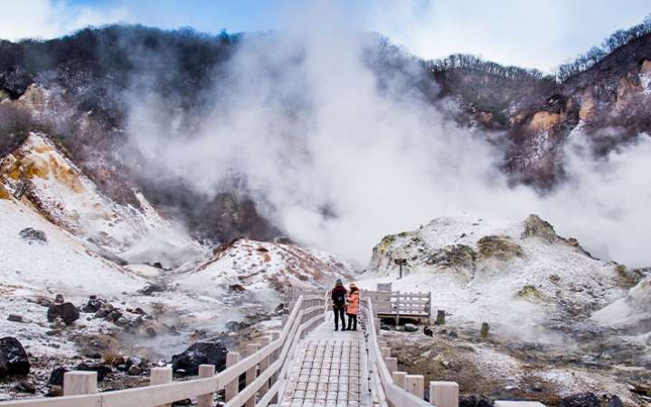 Steaming volcanic landscape of Noboribetsu Jigokudani (Hell Valley) in Hokkaido, Japan, with sulfur vents, rocky terrain, and hot springs surrounded by lush forest — a top geothermal attraction in Japan’s onsen town of Noboribetsu.