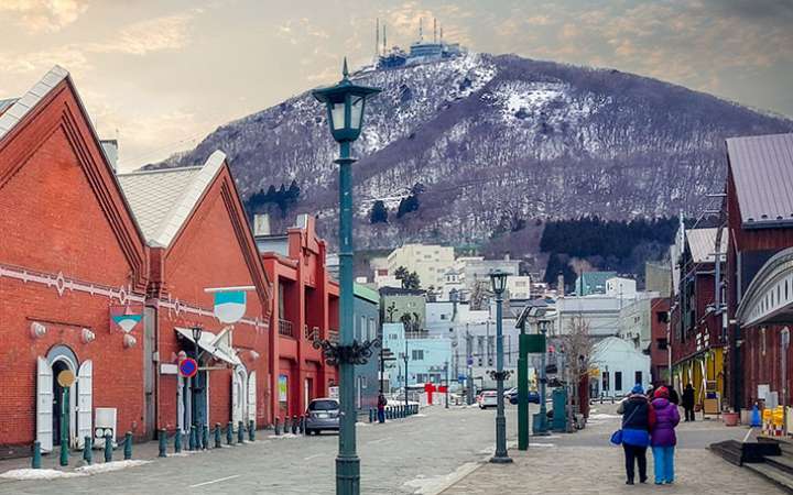 Historic red brick warehouses in Hakodate Bay, Hokkaido, Japan, reflecting on the water with a backdrop of Mount Hakodate — popular tourist spot featuring shops, cafes, and scenic views in a charming harbor district.
