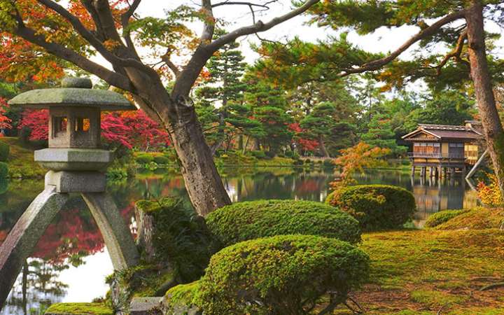 Scenic view of Kenrokuen Garden in Kanazawa, Japan, featuring traditional stone lanterns, manicured pine trees, a tranquil pond, and seasonal cherry blossoms under a clear blue sky