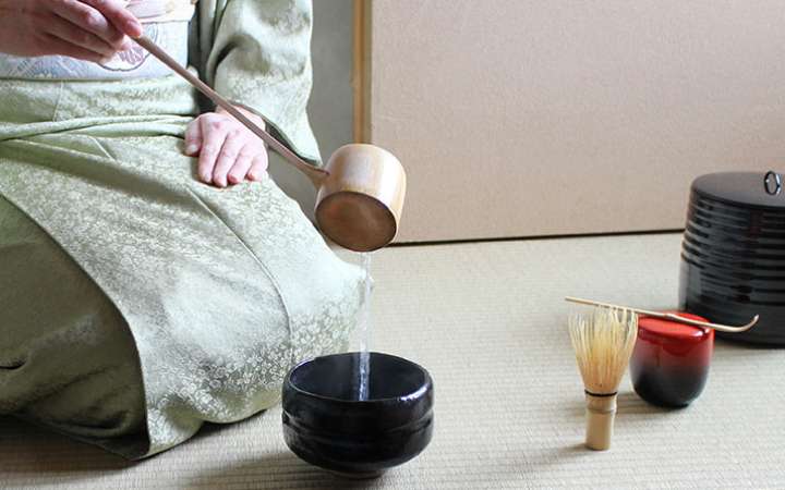 Traditional Japanese tea ceremony with a kimono-clad host preparing matcha in a tatami room, featuring a tea bowl, bamboo whisk (chasen), and seasonal flower arrangement.